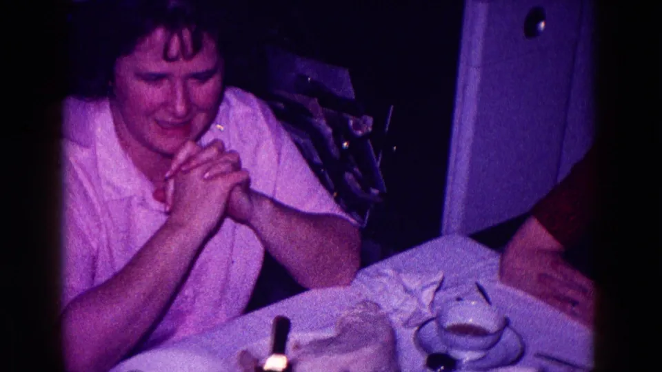 A younger adult woman in a light shirt sits with clasped hands at a table in the same 1968 Minnesota home interior.