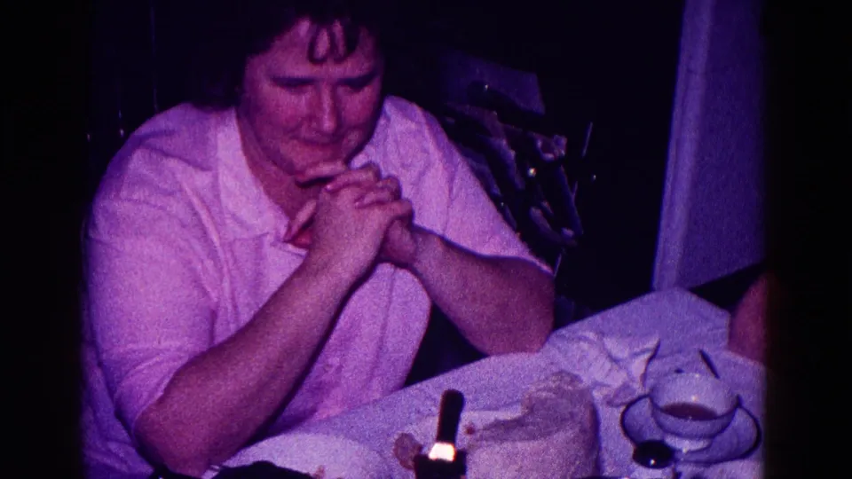 1968 Minnesota home-movie frame of a woman in a light shirt clasping her hands at a dinner table with teacups and dessert visible.