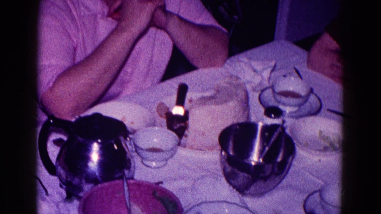 Tabletop with clasped hands, metal teapot, cups, saucers, and cake remnants.