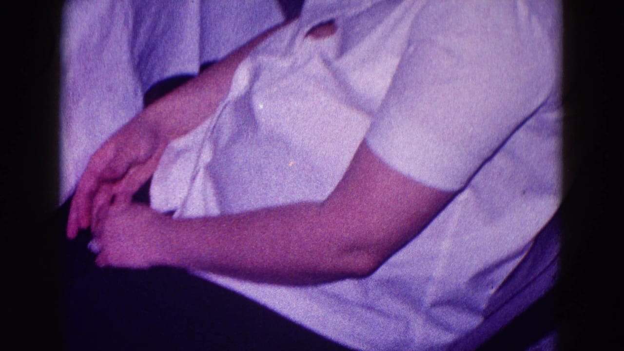 1968 Minnesota home-movie frame showing a seated woman in white clothing, cropped at the face, with hands in her lap.