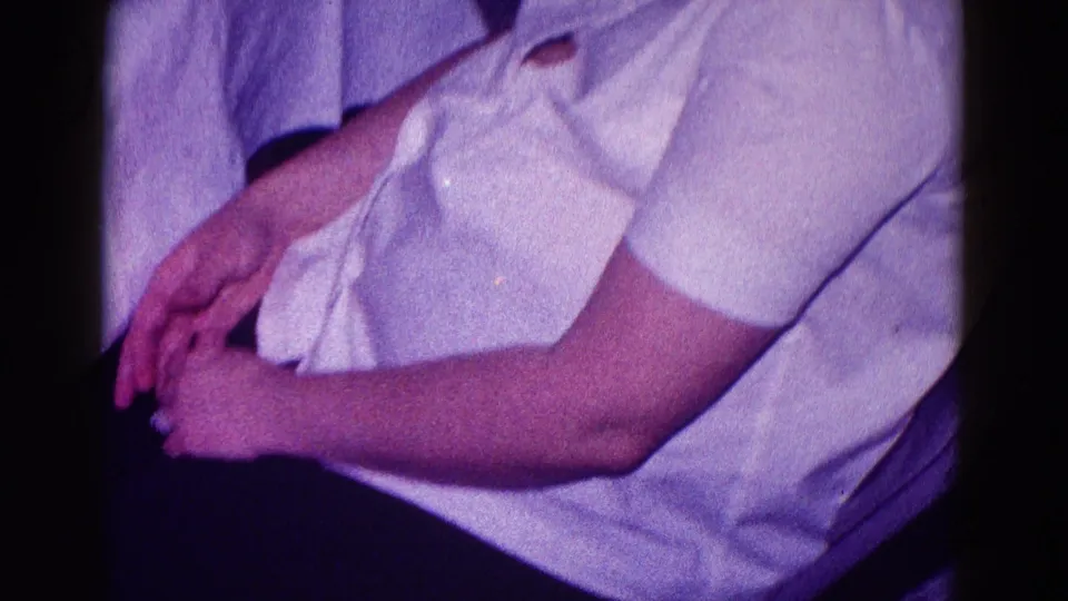1968 Minnesota home-movie frame showing a seated woman in white clothing, cropped at the face, with hands in her lap.