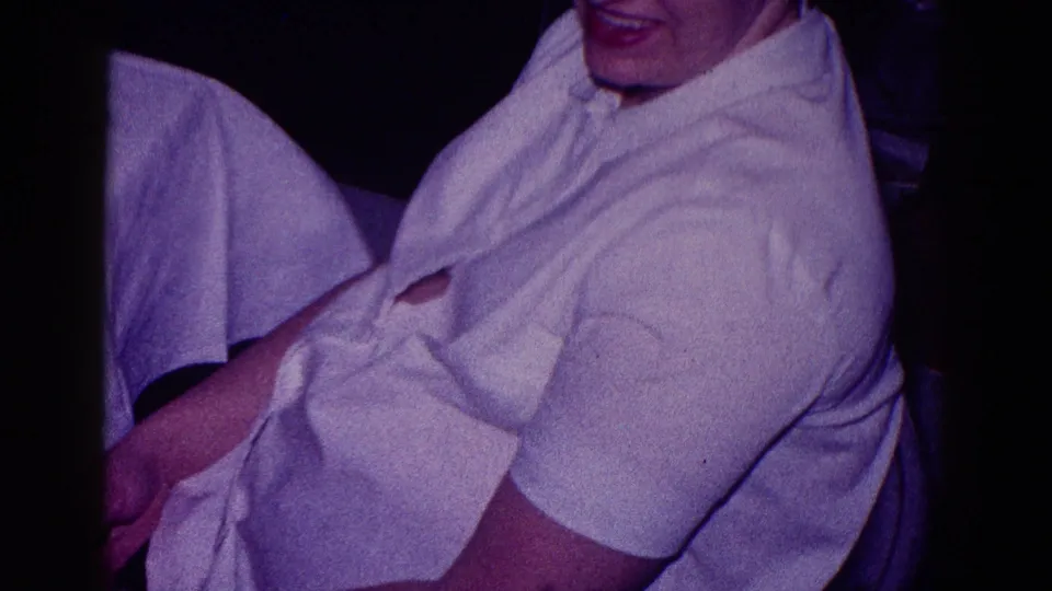 1968 Minnesota 8mm frame of a seated woman in a white blouse, partly smiling or speaking in a dim interior.