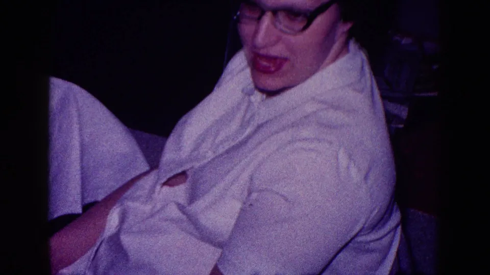 1968 Minnesota home-movie frame showing a seated older woman with glasses and a white blouse, viewed from below indoors.