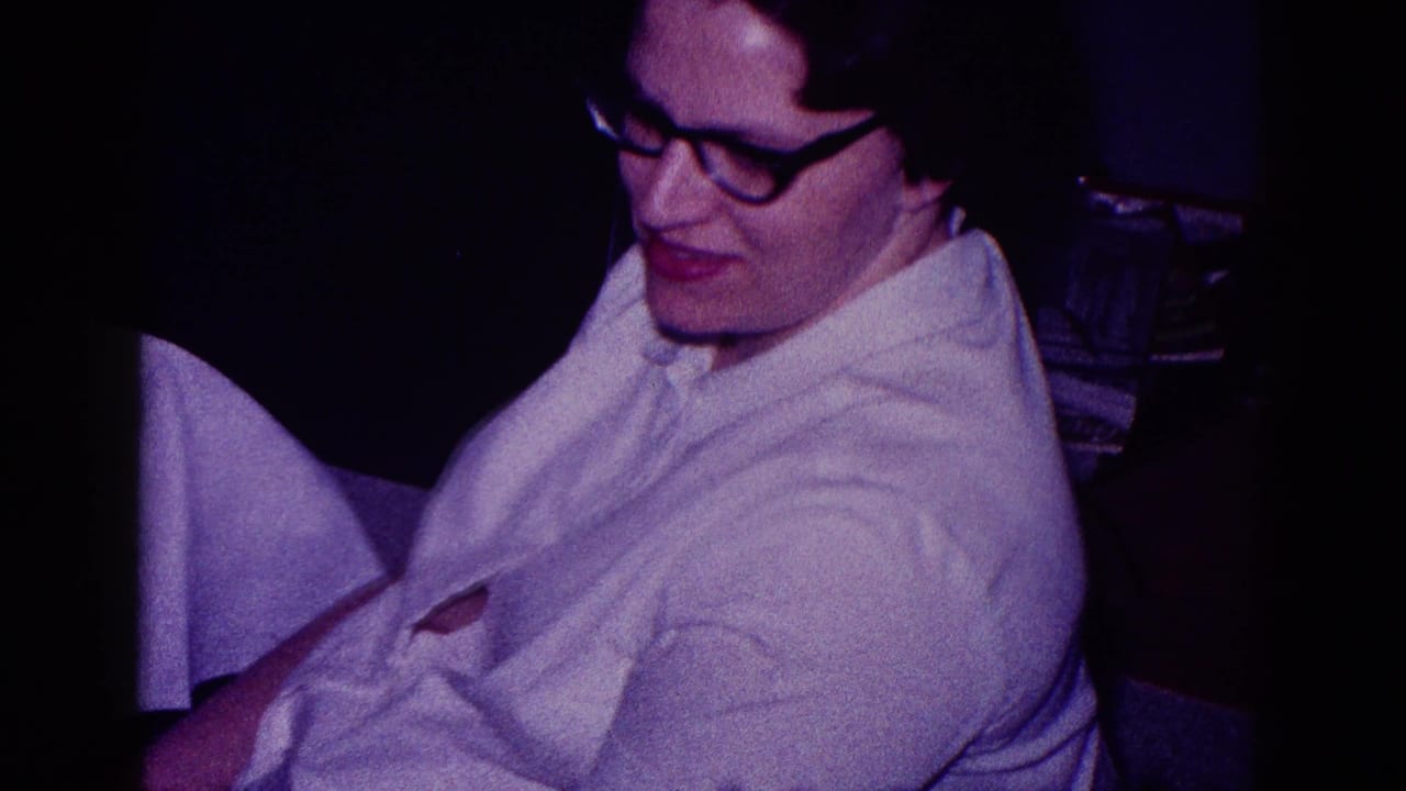 Woman in glasses and a white shirt seated in a dim interior.