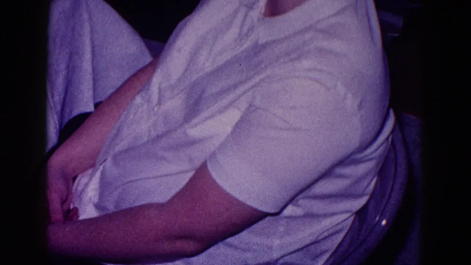 Side view of a seated adult in a white shirt within a dim private interior, captured with soft 8mm color and grain.