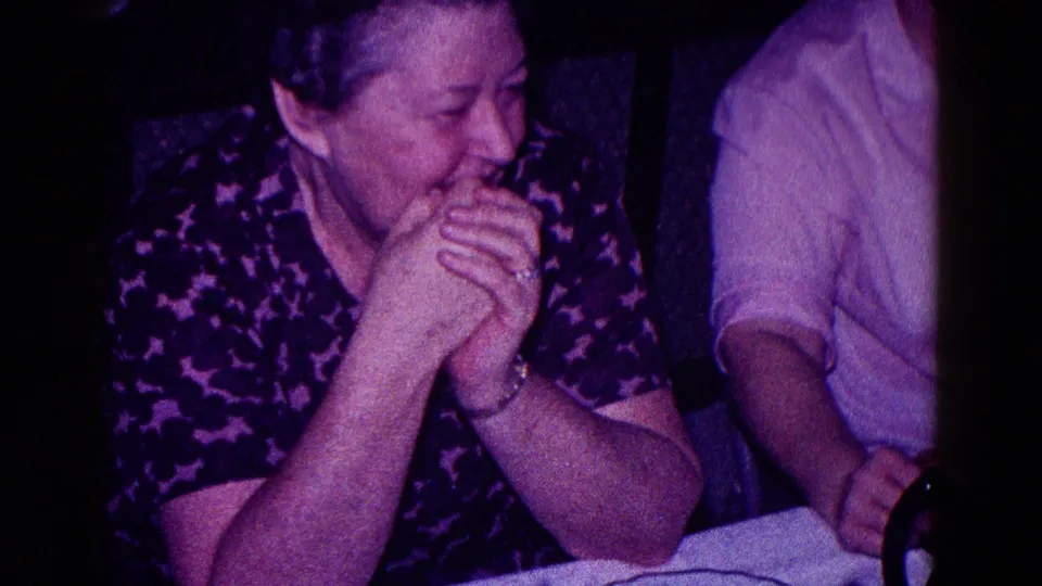 Adult woman in a patterned blouse laughs with clasped hands beside other cropped seated figures in a 1968 Minnesota home movie.