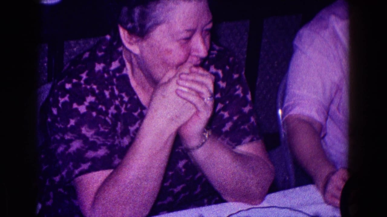 In 1968 Minnesota home-movie footage, an older woman in a dark patterned top smiles with clasped hands beside another seated person.