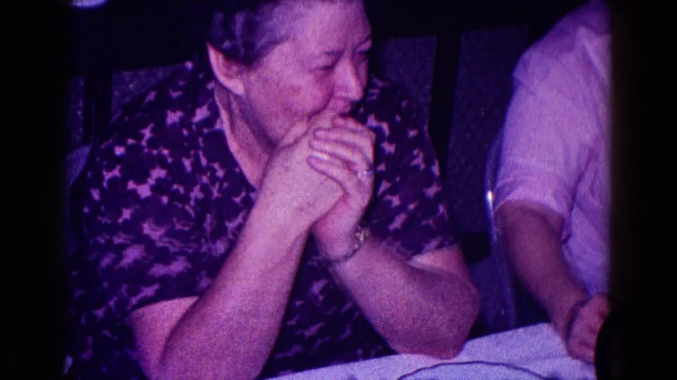 In 1968 Minnesota home-movie footage, an older woman in a dark patterned top smiles with clasped hands beside another seated person.