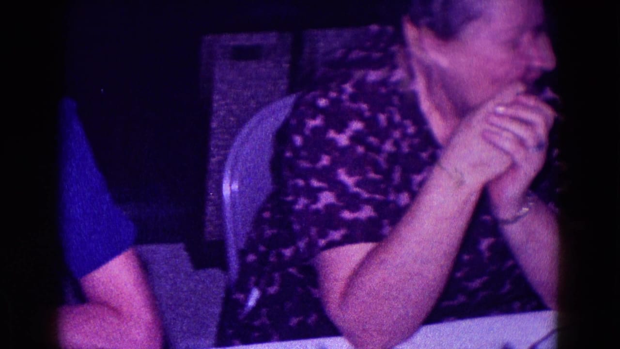 An older woman in a dark patterned top turns toward someone off frame while seated at a table in the Minnesota 1968 footage.