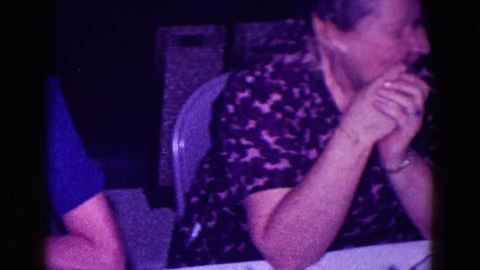 An older woman in a dark patterned top turns toward someone off frame while seated at a table in the Minnesota 1968 footage.