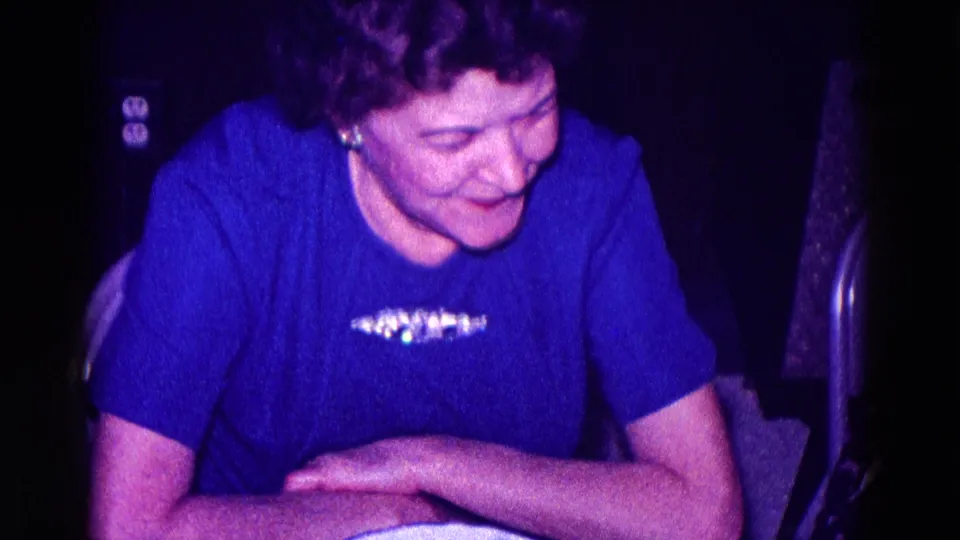 An older woman wearing a royal blue blouse with a decorative brooch smiles downward in this 1968 Minnesota indoor home-movie frame.
