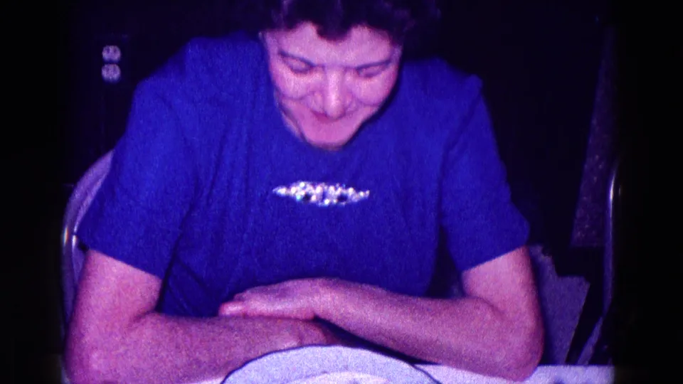 The woman in the royal blue blouse and brooch looks down with folded arms at the table in the 1968 Minnesota home-movie scene.