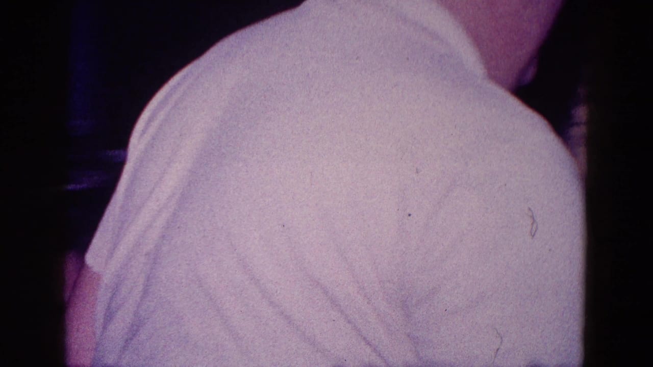 1968 Minnesota 8mm frame showing the back and shoulder of a man in a white polo shirt inside a dark private room.