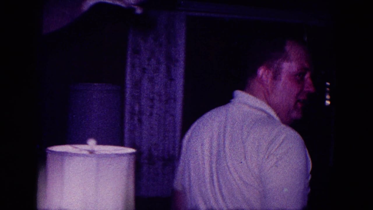 Adult man in a white polo shirt turns inside a dim Minnesota home interior, with a lampshade and curtains visible.