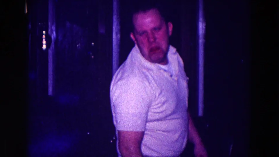 The man faces toward the camera with a blurred expression in a dim 1968 Minnesota home-movie interior.