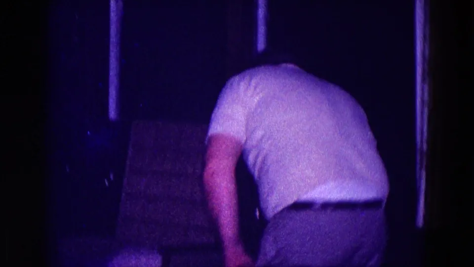 Adult man in a white polo shirt bends forward inside the dim private room, with curtains and dark wall surfaces nearby.