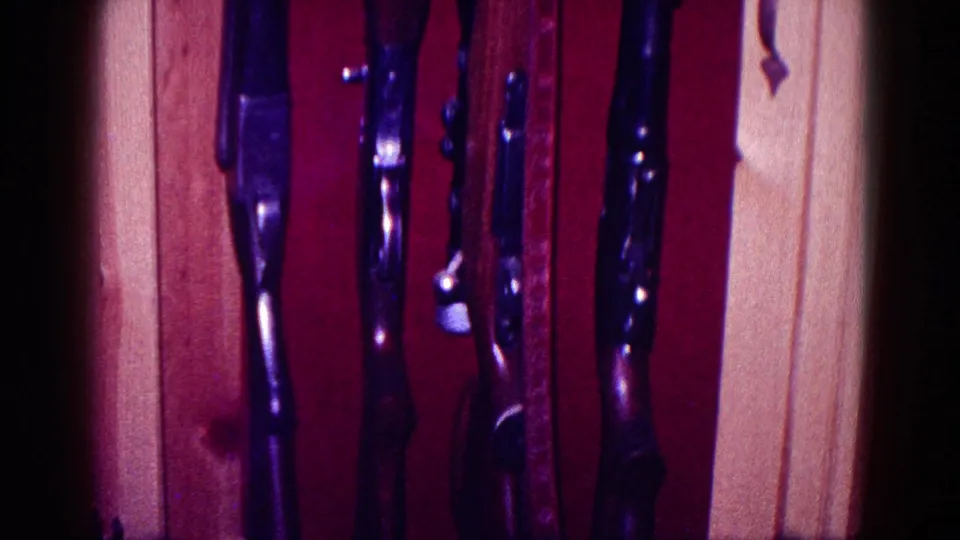 Close 8mm view of multiple rifles standing in a wooden display cabinet, with red fabric backing and soft focus.