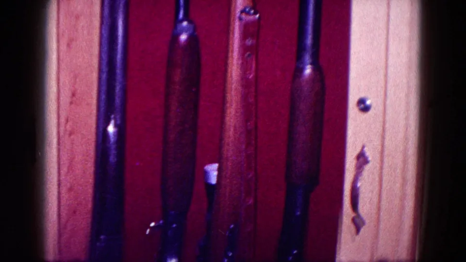Home-movie frame showing rifle stocks inside a red-lined wooden cabinet beside a light wood side panel and small handle.