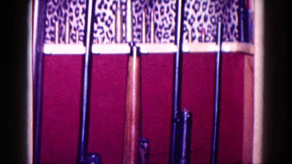 1968 Minnesota 8mm frame showing upright long guns beneath patterned trim and red fabric inside a wooden cabinet.