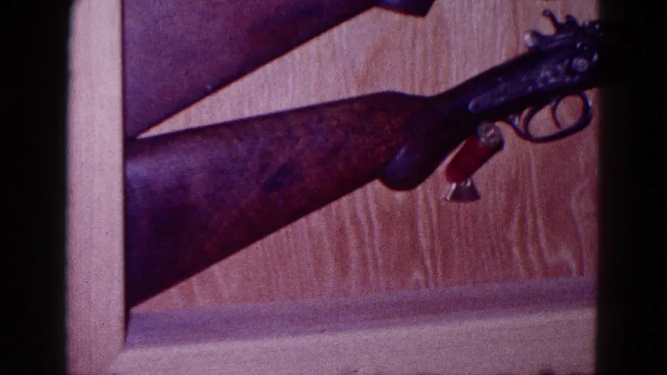1968 Minnesota home-movie frame showing a wood-stock shotgun, trigger guard, and red shell against wood paneling.