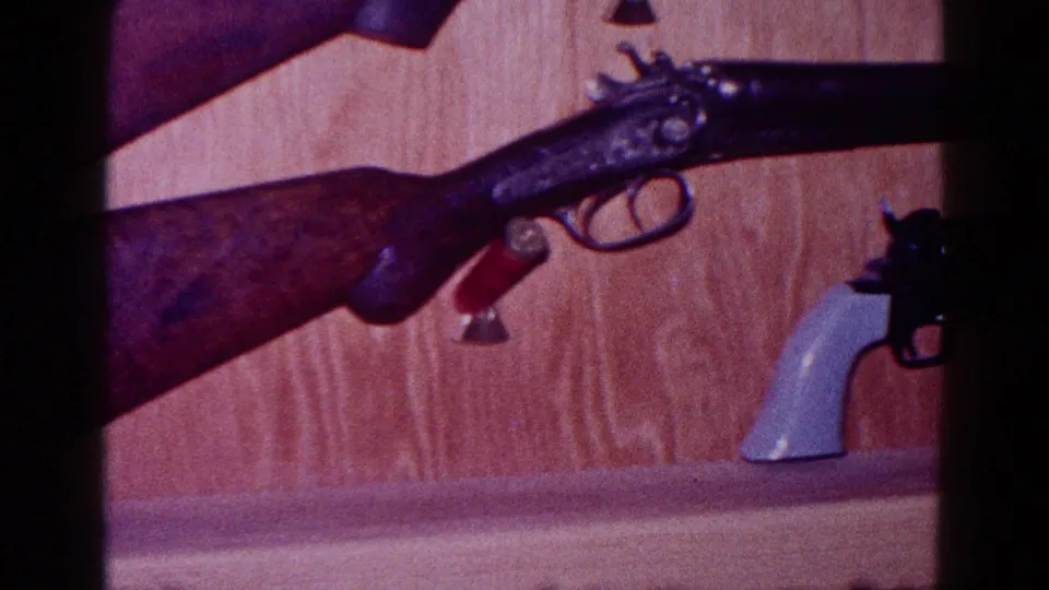 1968 Minnesota home-movie frame with a mounted long gun above a partially visible light-handled revolver.