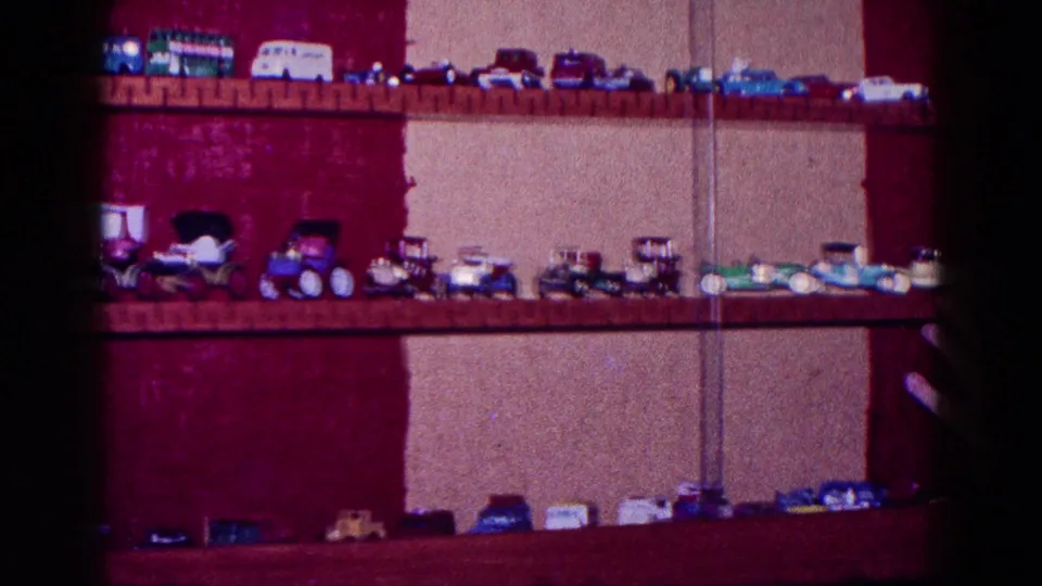 1968 Minnesota home-movie frame with miniature cars and trucks displayed across wooden shelves against red and beige walls.