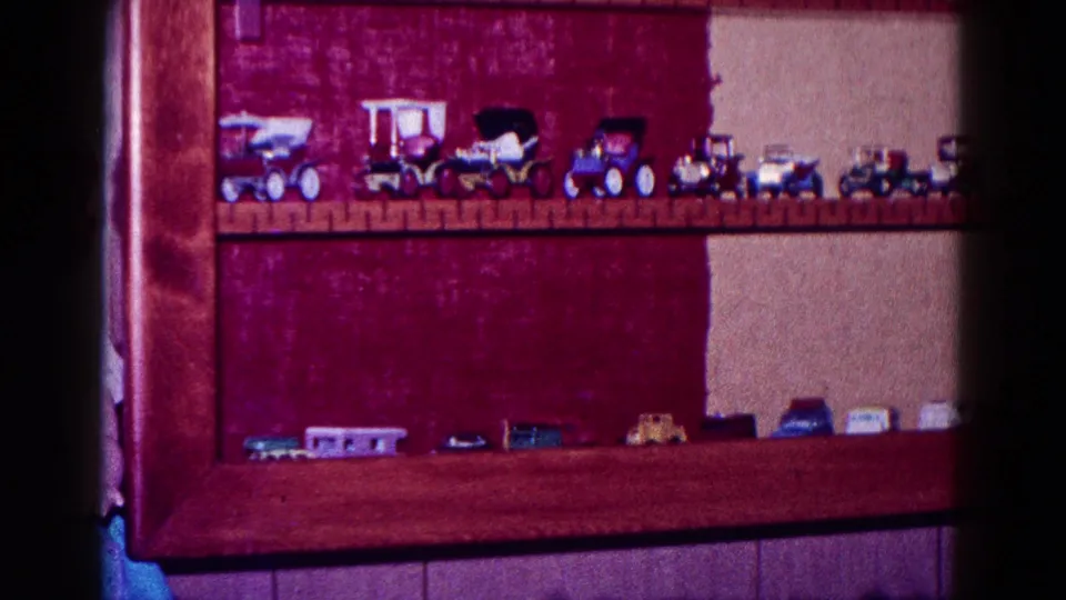 1968 Minnesota home-movie frame of a toy vehicle collection spread across wooden shelves in a domestic room.