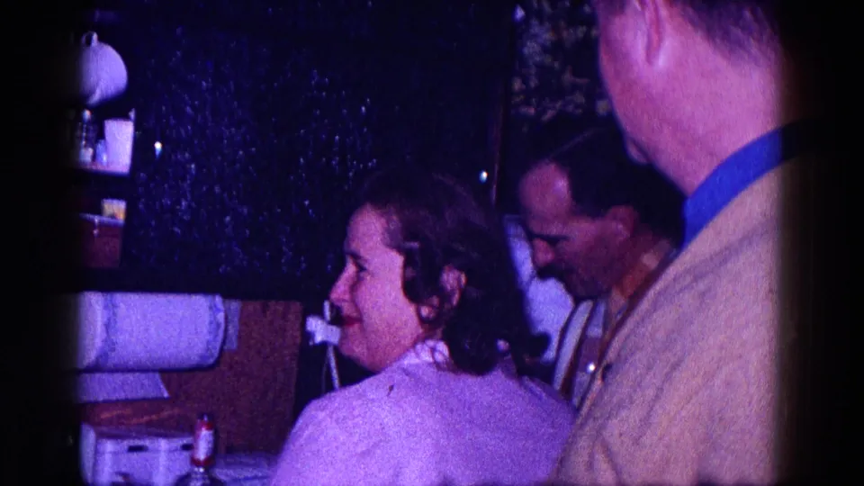 1968 Minnesota home-movie frame of adults standing close together and smiling near kitchen shelves in a private interior.