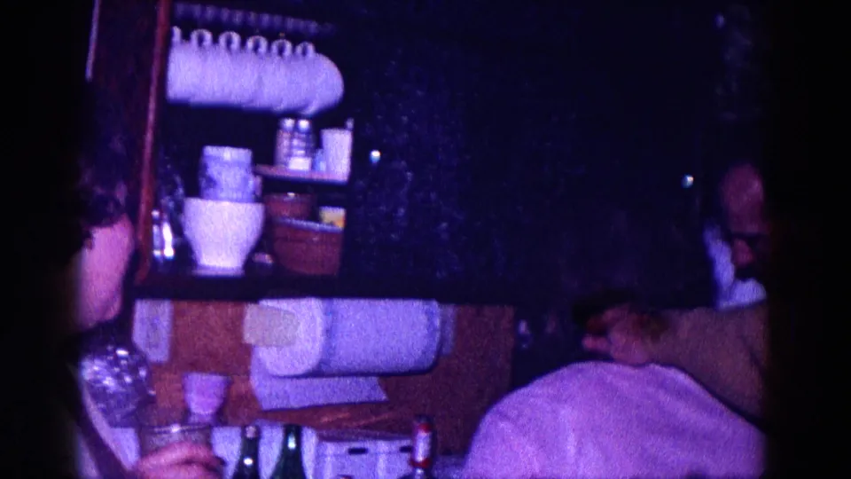 1968 Minnesota 8mm frame showing adults in a dim private kitchen area with shelves, dishes, cups, and strong purple film color.