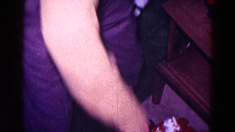 1968 Minnesota home-movie frame with a man's arm reaching near a wrapped Christmas present beside a dark chair.