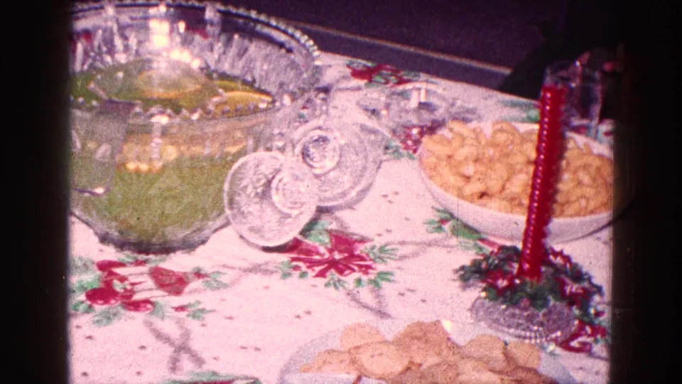 Holiday table spread with green punch, red taper candle in a wreath holder, snack bowl, and cookies in 1968 Minnesota home footage.