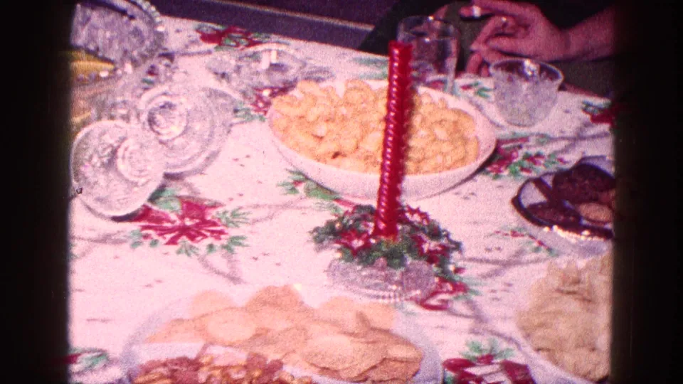 Christmas party table with cookies, a snack bowl, red candle, glassware, and adults' hands partly visible in the Minnesota home-movie frame.