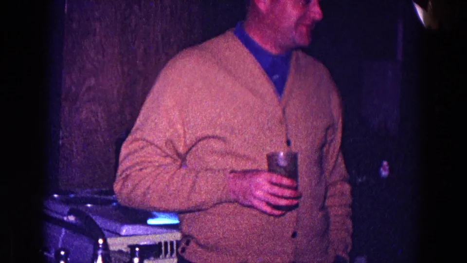 A man in a beige cardigan holds a drink while standing in a dim private interior during a 1968 Minnesota party.