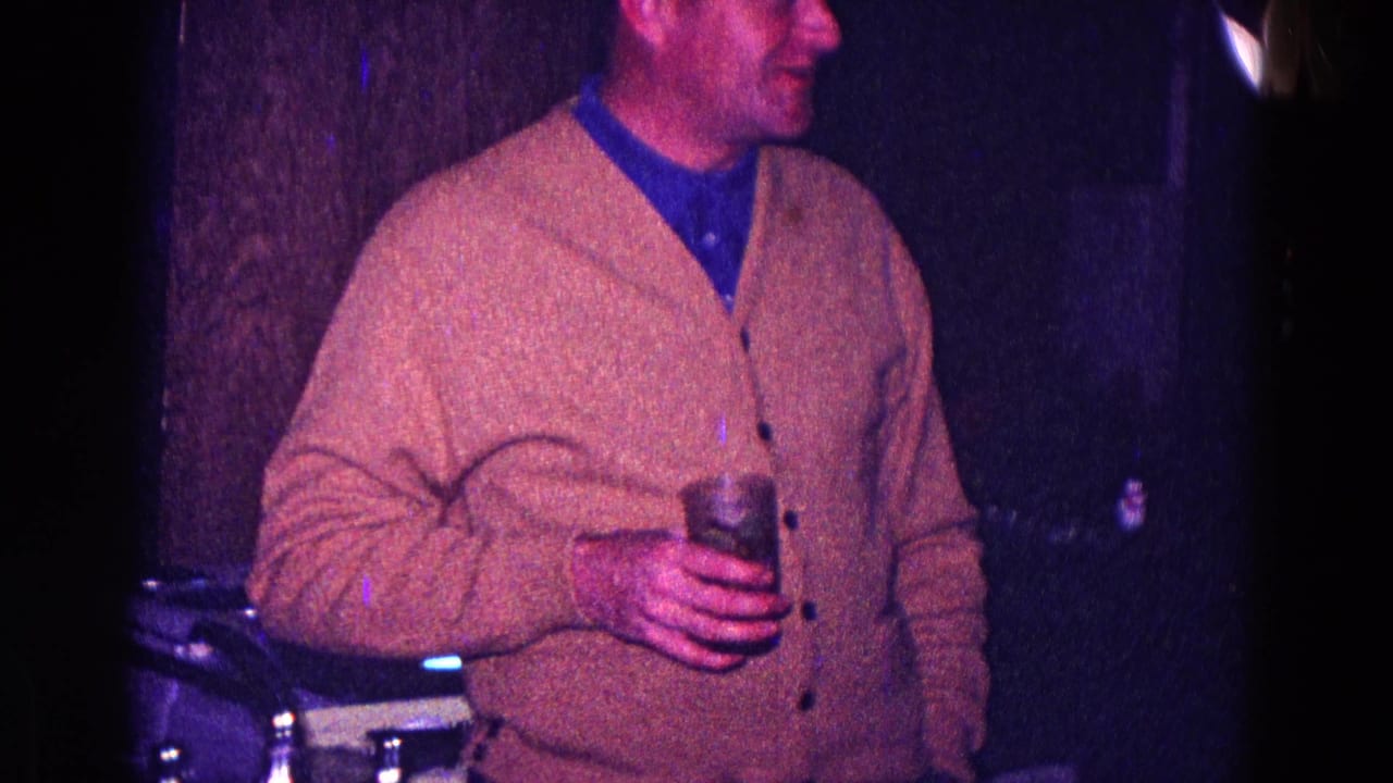 In 1968 Minnesota home-movie footage, a man in a tan cardigan and blue shirt holds a drink in a dark, color-shifted room.