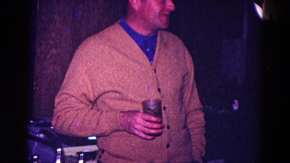 In 1968 Minnesota home-movie footage, a man in a tan cardigan and blue shirt holds a drink in a dark, color-shifted room.