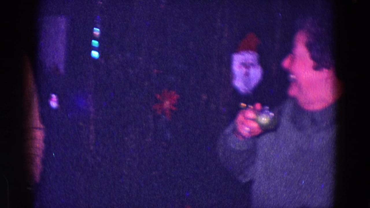 A woman in a gray sweater holds a drink and smiles near a Santa face ornament and colored Christmas lights in a 1968 Minnesota home movie.