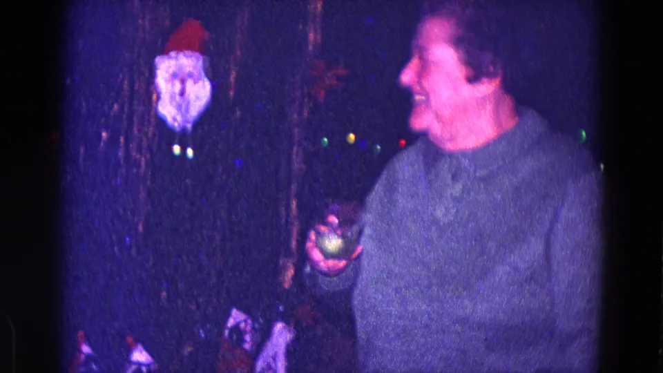 The woman laughs with a drink in hand beside a Santa face ornament, colored lights, and dark interior decor in the 1968 Minnesota holiday scene.