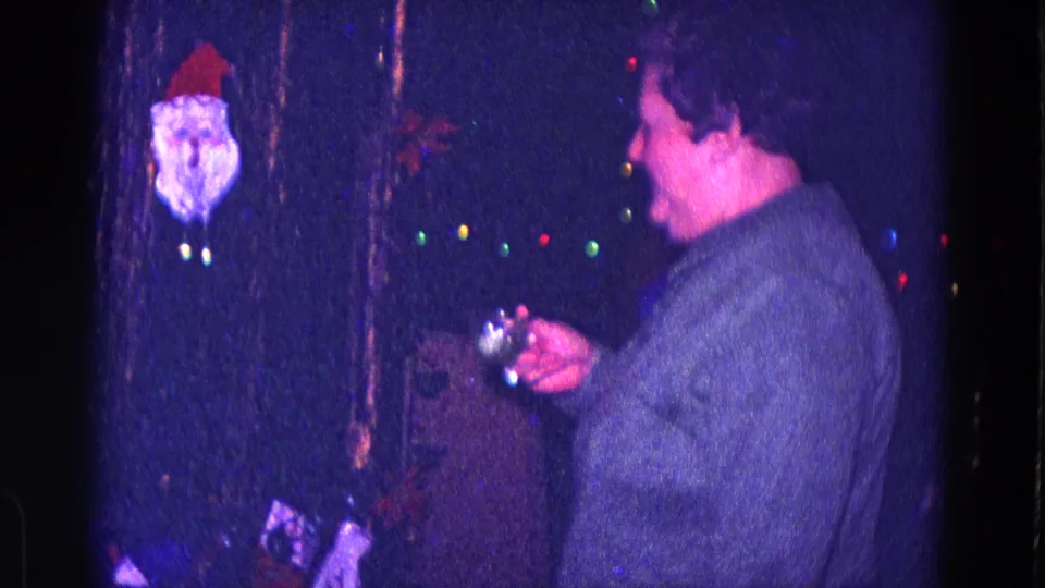 A woman in a gray sweater turns toward Christmas lights and a Santa ornament while holding a small glass in 1968 Minnesota home-movie footage.