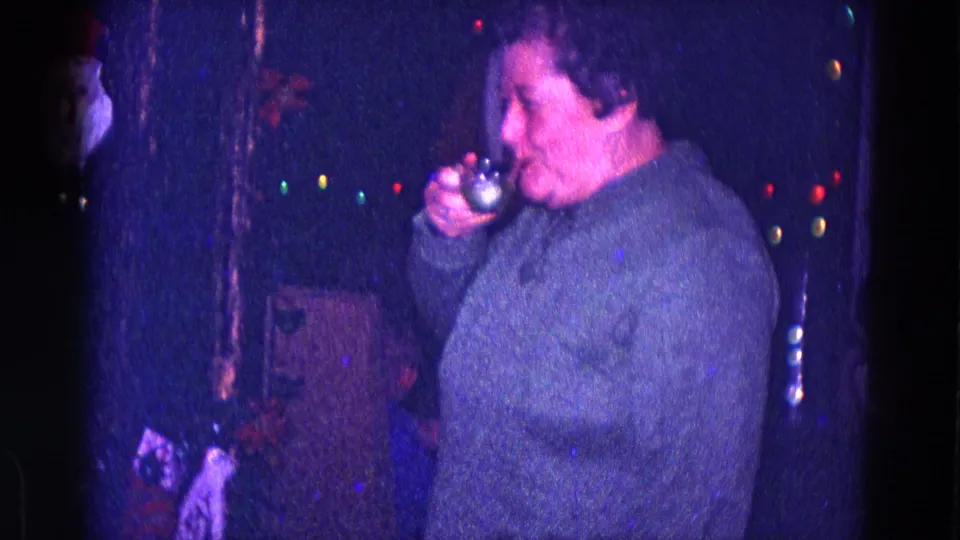 A woman drinks from a small glass in dim 1968 Minnesota Christmas home-movie footage with colored lights behind her.