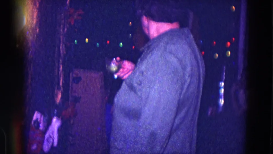 The woman turns partly away while holding her glass, with colored Christmas lights and holiday decor visible in the dark interior.