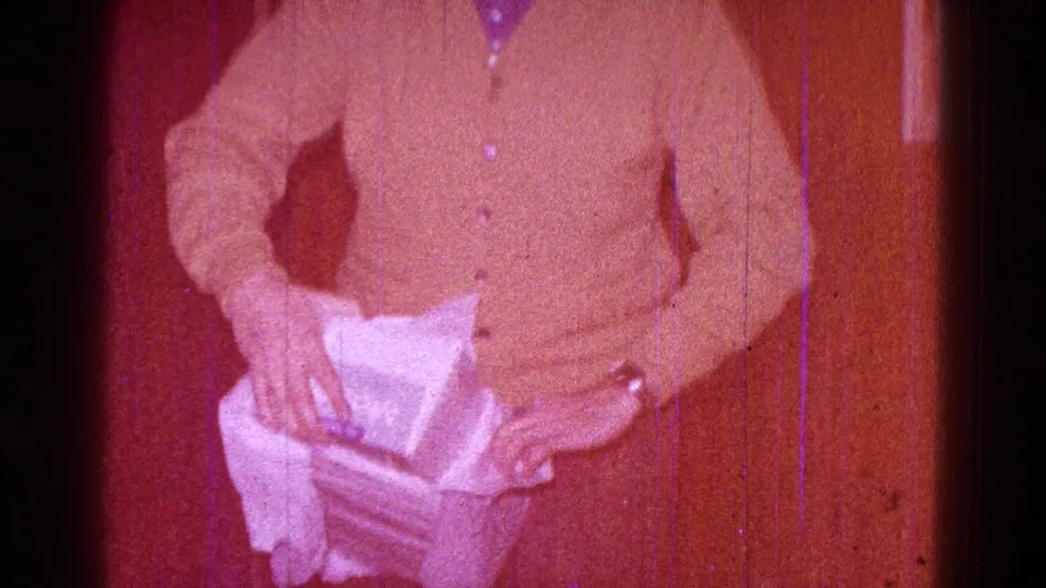 1968 Minnesota home-movie frame of a man in a brown cardigan handling a wrapped package inside a private interior.