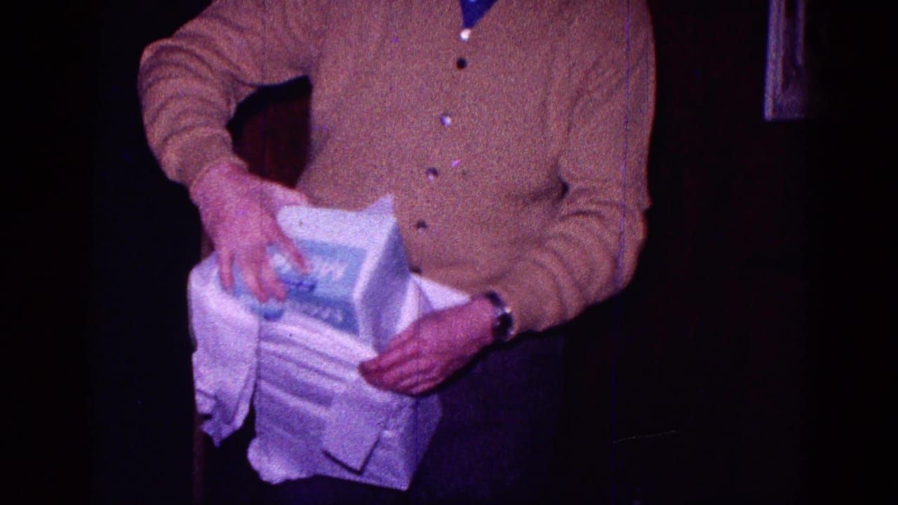 In a dim Minnesota home interior from 1968 metadata, a man in a brown cardigan pulls paper from a large white gift box.