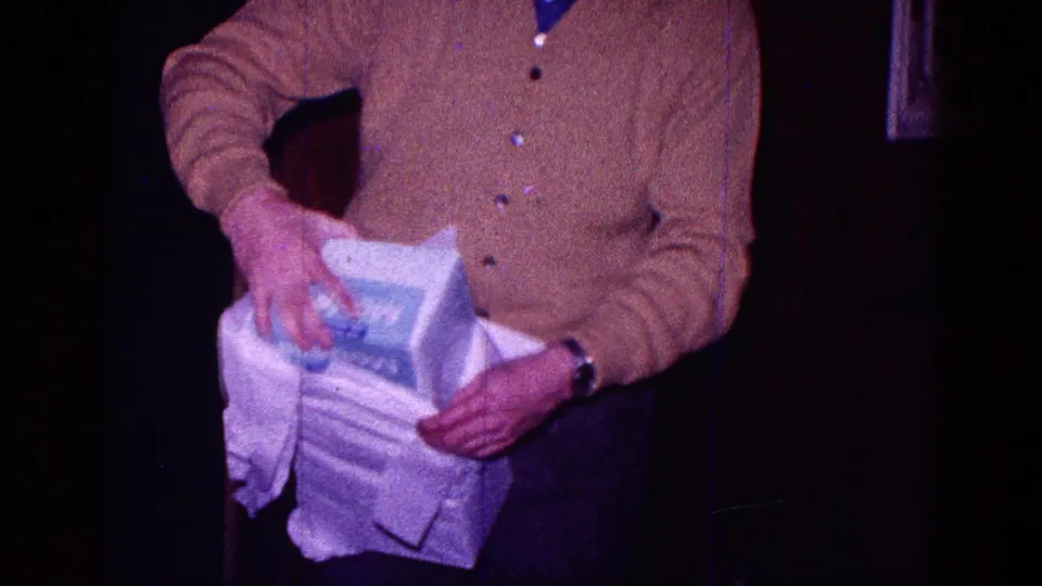 In a dim Minnesota home interior from 1968 metadata, a man in a brown cardigan pulls paper from a large white gift box.
