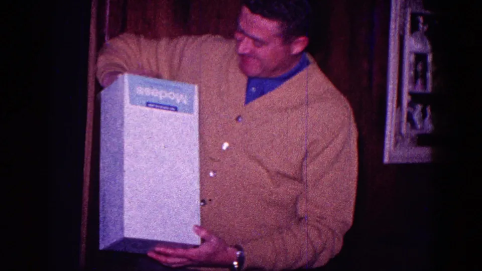 The man looks at the large white box after unwrapping it, with a blue product label visible but not clearly identifiable.