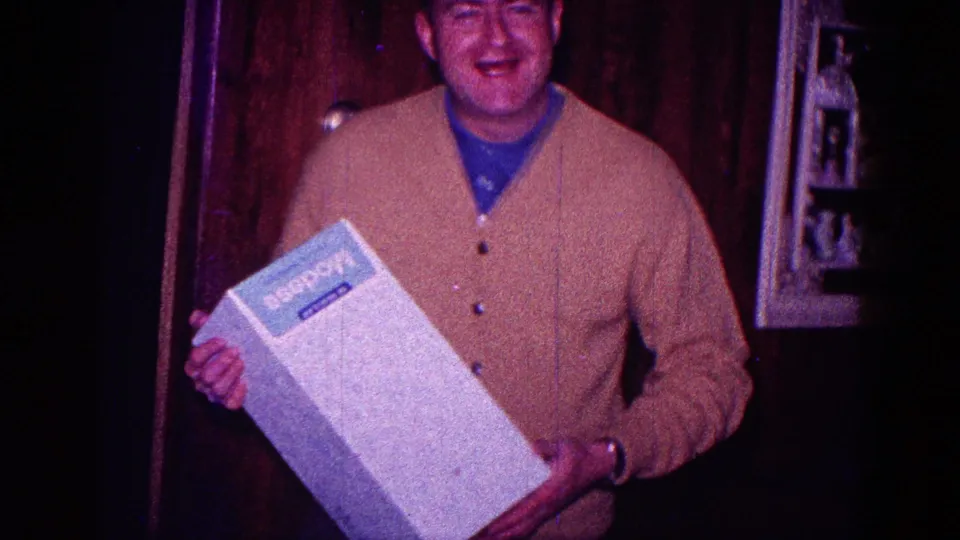 A smiling man in a brown cardigan presents the large white boxed gift toward the camera in a dim 1968 Minnesota home interior.