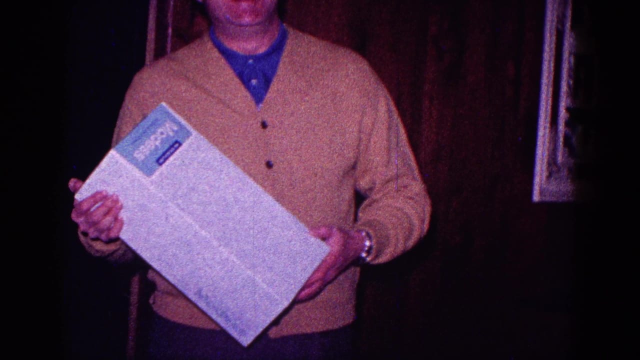 Man in a beige cardigan holds a white box with a blue label in a dark wood-paneled room.