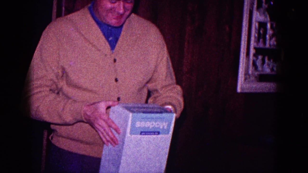 Man grips the white box at his waist while standing against dark wood paneling.