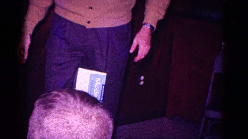 The man places the box between his knees in the wood-paneled room, with another person's head visible low in the foreground.