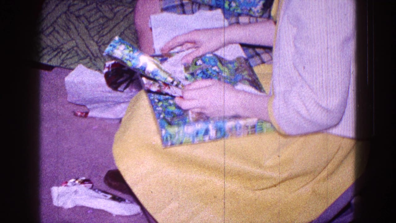 In 1968 Minnesota home-movie footage, a child in a yellow dress and pale sweater holds a floral-wrapped present indoors.
