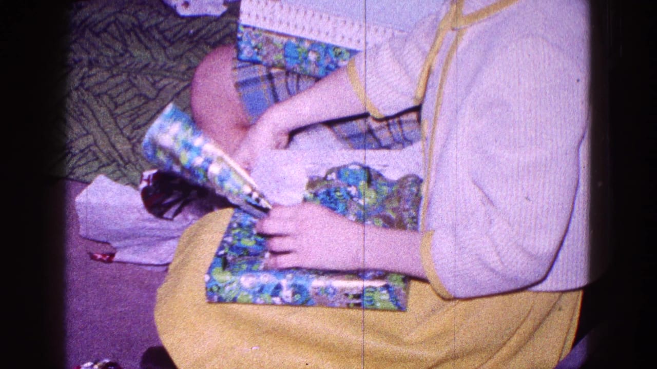 Child tears blue-green wrapping paper from a present while seated indoors.